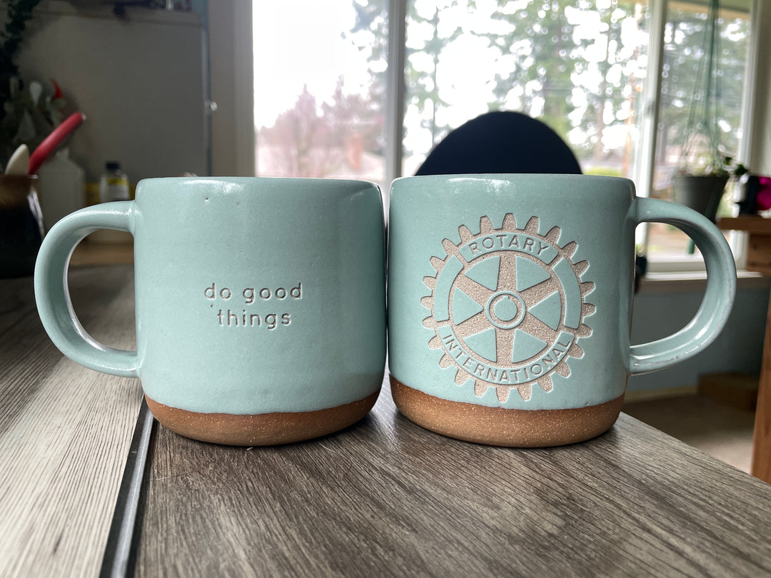 "do good things" engraved on a light blue mug, and the Rotary International logo engraved onto another blue mug sitting next to it.