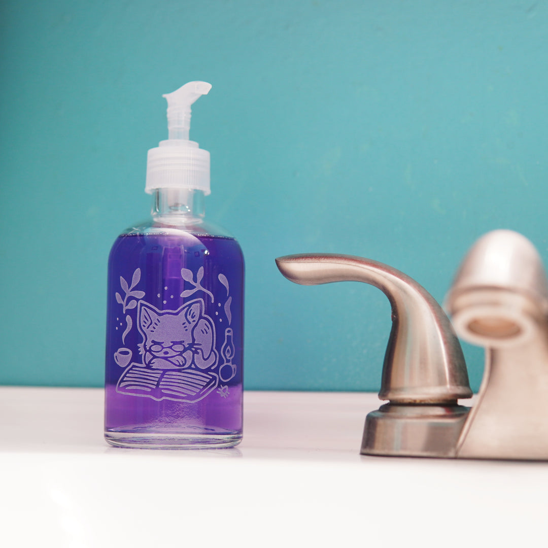 glass pump bottle soap dispenser full of purple soap next to a bathroom faucet, with a cat sleeping on a book engraved on it