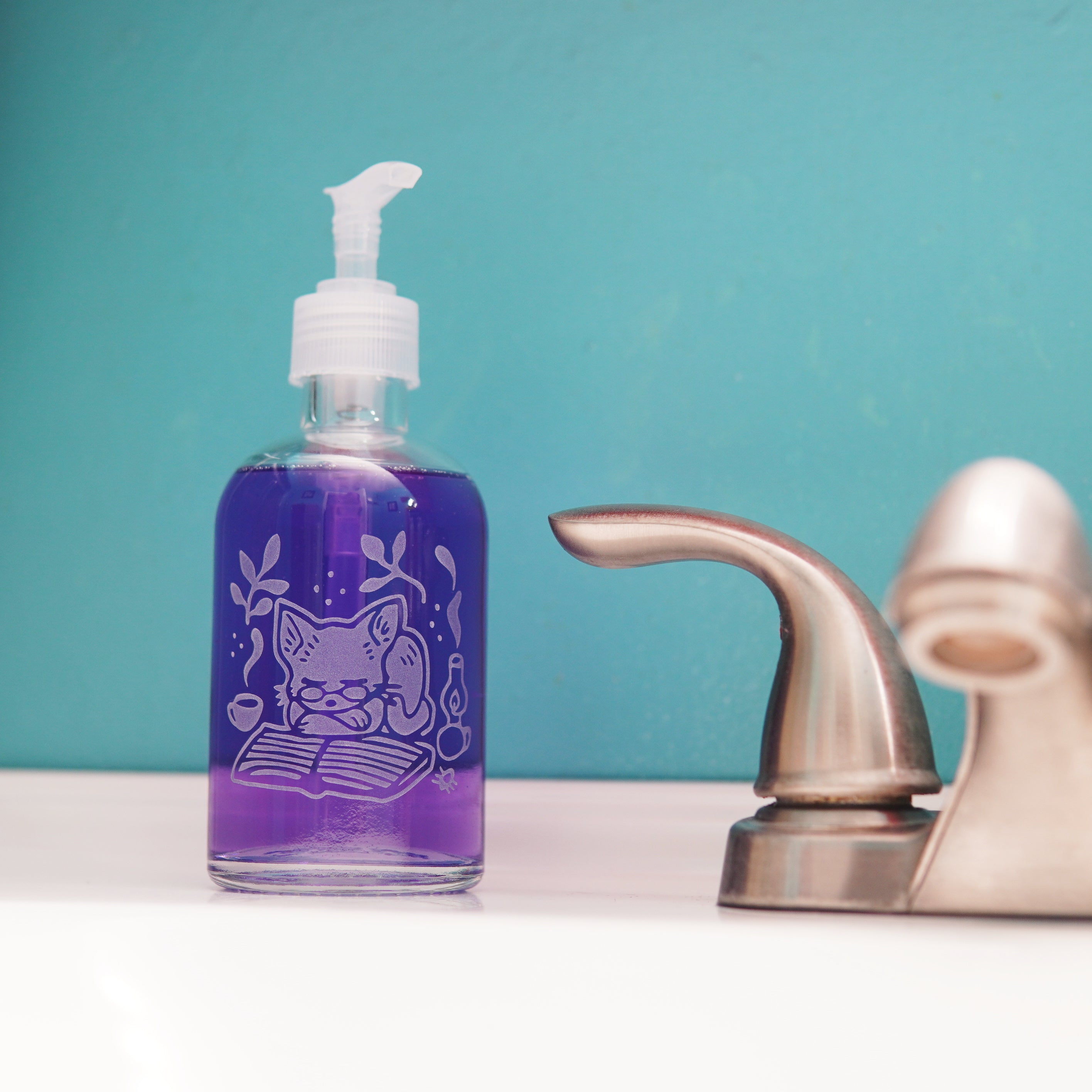 glass pump bottle soap dispenser full of purple soap next to a bathroom faucet, with a cat sleeping on a book engraved on it
