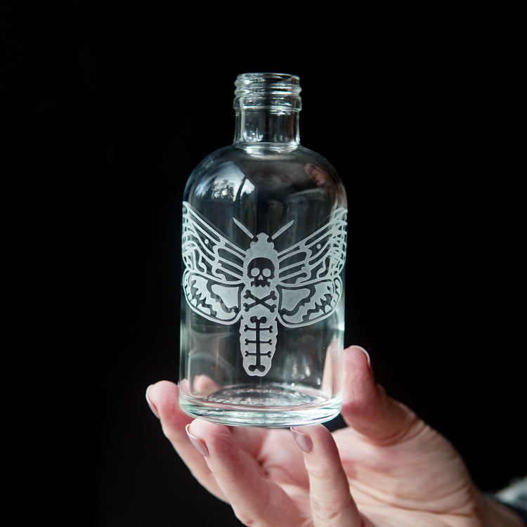 glass bottle engraved with a deaths head hawkmoth, held by a hand in front of a black background
