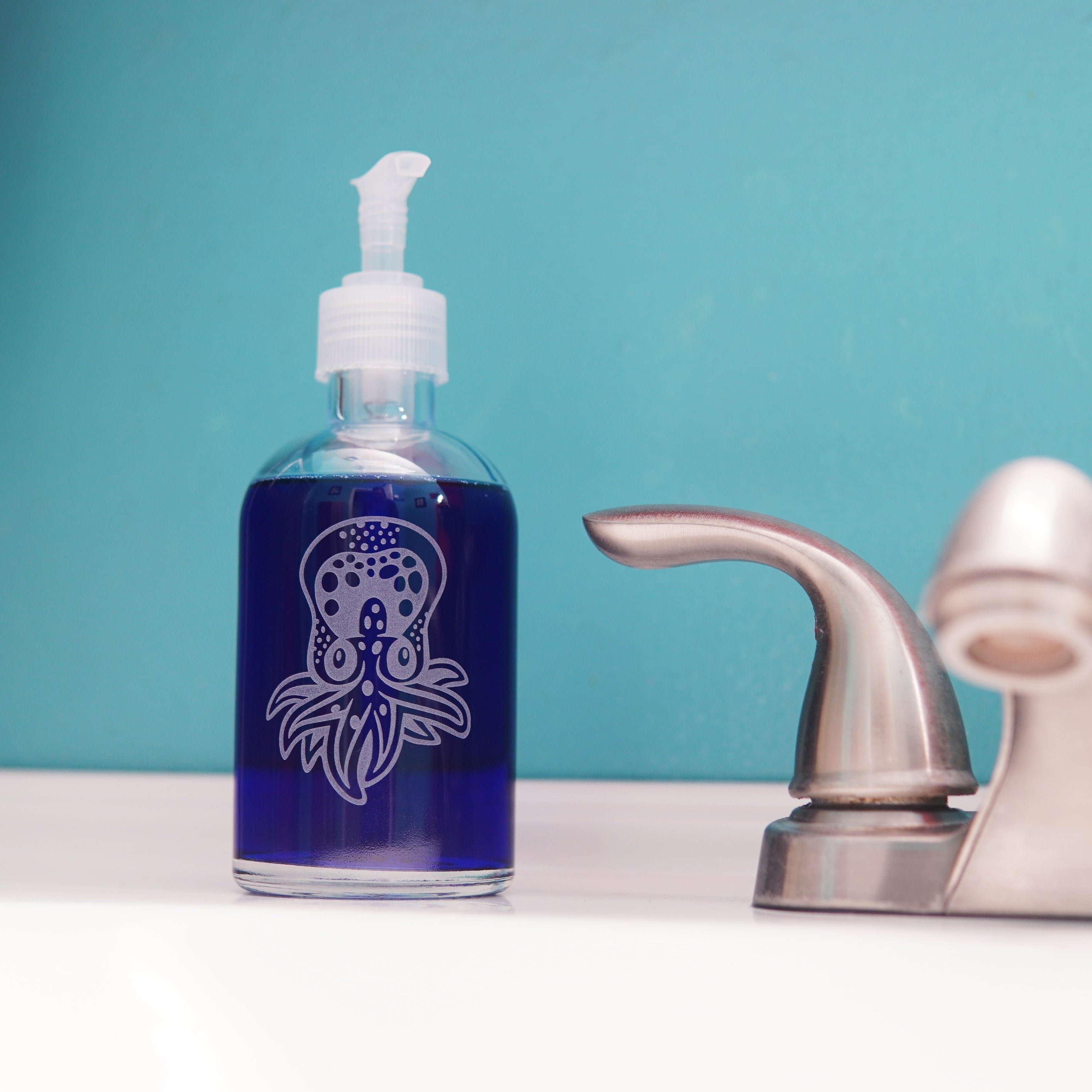 glass pump bottle soap dispenser full of dark blue soap next to a bathroom faucet, with an octopus engraved on it