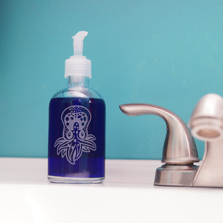 glass pump bottle soap dispenser full of dark blue soap next to a bathroom faucet, with an octopus engraved on it