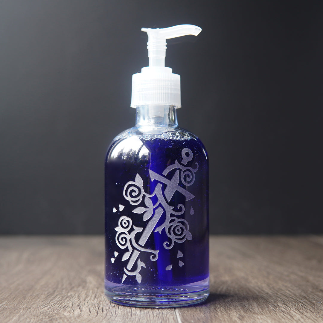 glass soap dispenser engraved with a sword wrapped with a rose vine. the pump bottle is filled with dark blue liquid soap