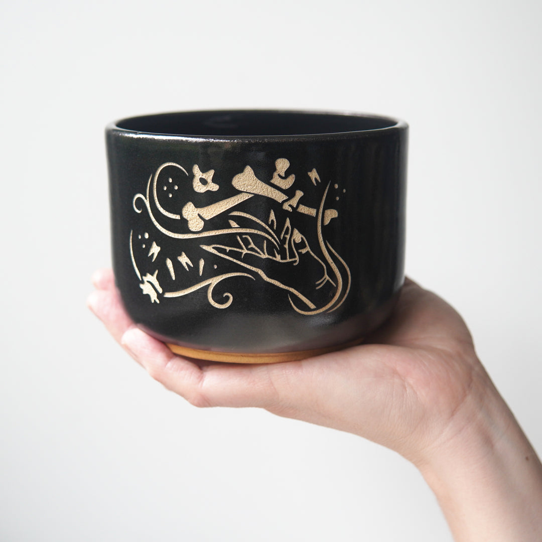 Medium sized black pottery bowl held by a hand. The bowl has an engraved graphic of a hand levitating human bones and teeth with swirls of magic