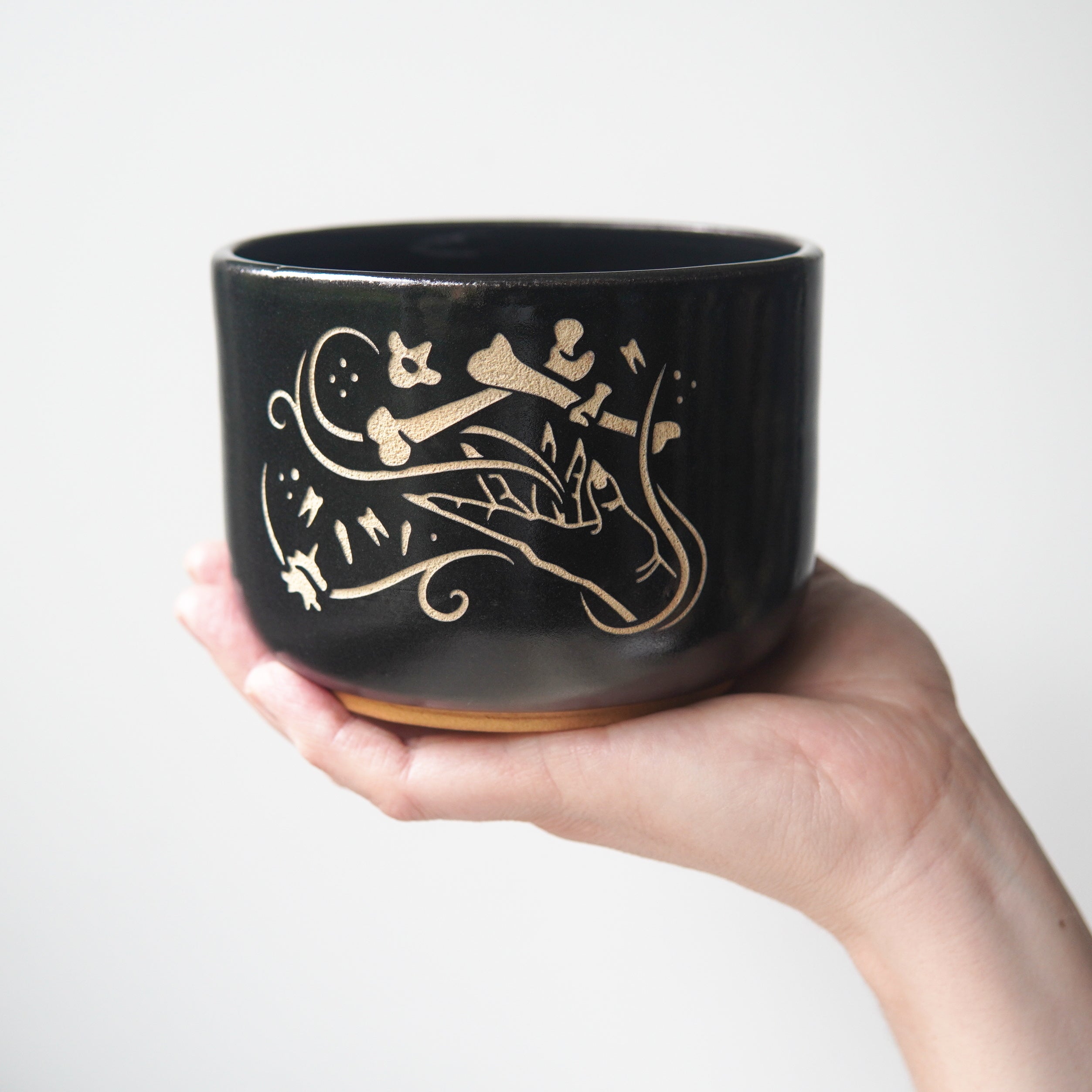 Medium sized black pottery bowl held by a hand. The bowl has an engraved graphic of a hand levitating human bones and teeth with swirls of magic