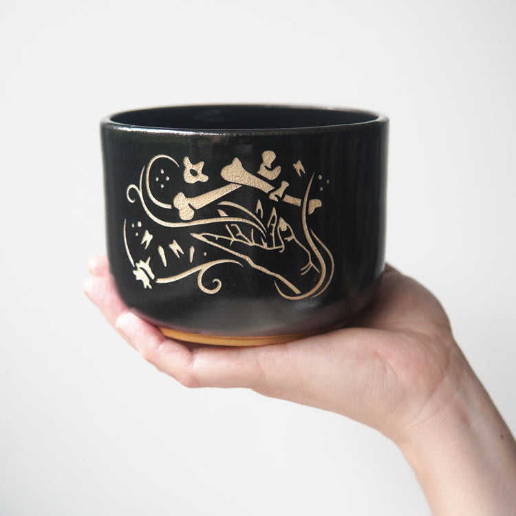 Medium sized black pottery bowl held by a hand. The bowl has an engraved graphic of a hand levitating human bones and teeth with swirls of magic