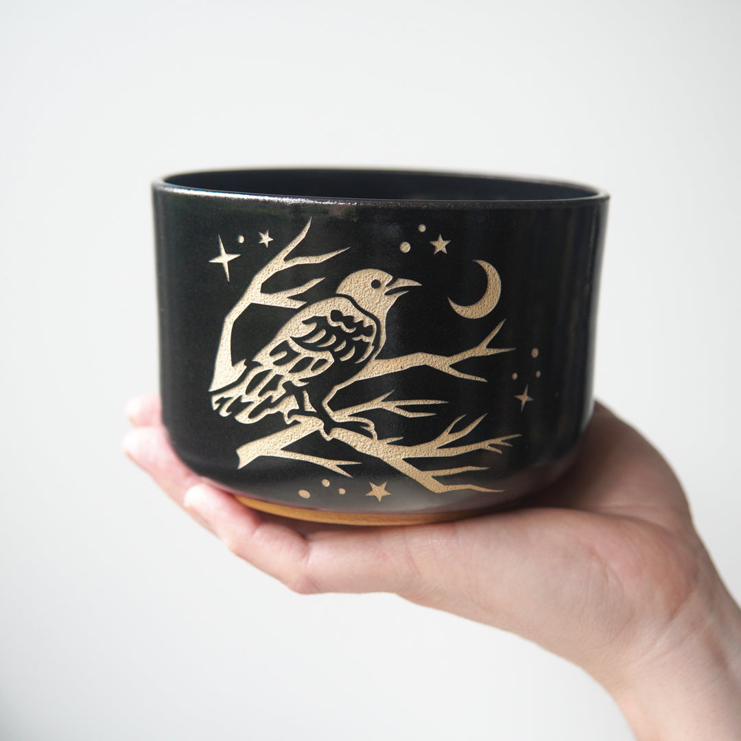 A black pottery bowl, held up by a hand. A crow perched in a tree looking at the crescent moon and stars is engraved on the bowl.