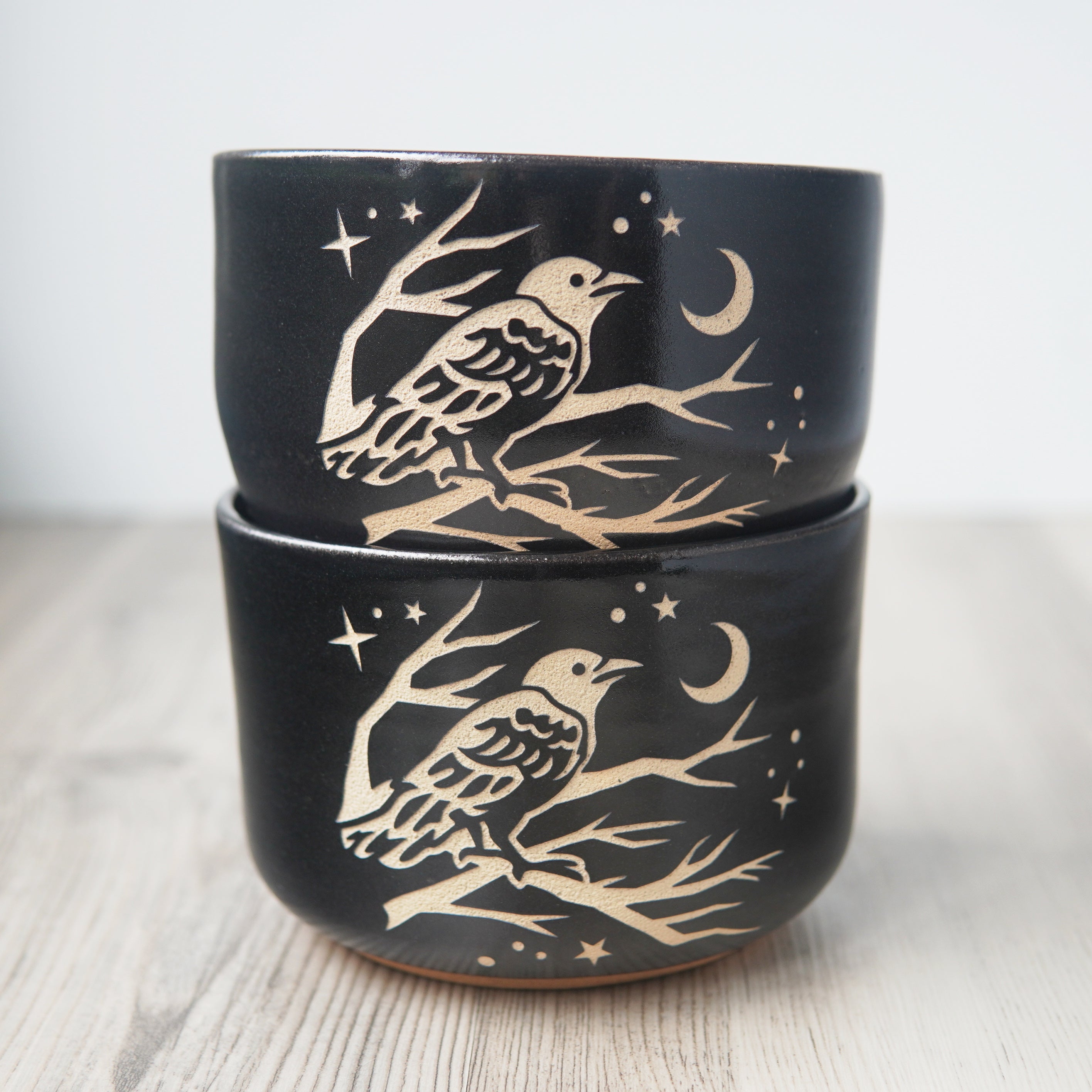 Two black pottery bowls, stacked on each other. A crow perched in a tree looking at the crescent moon and stars is engraved on the bowls.