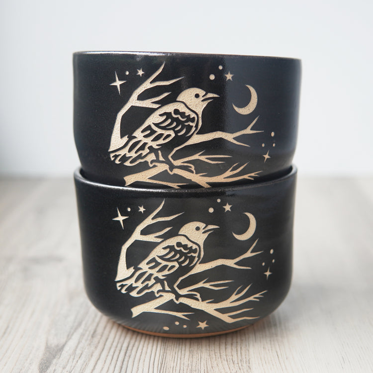 Two black pottery bowls, stacked on each other. A crow perched in a tree looking at the crescent moon and stars is engraved on the bowls.