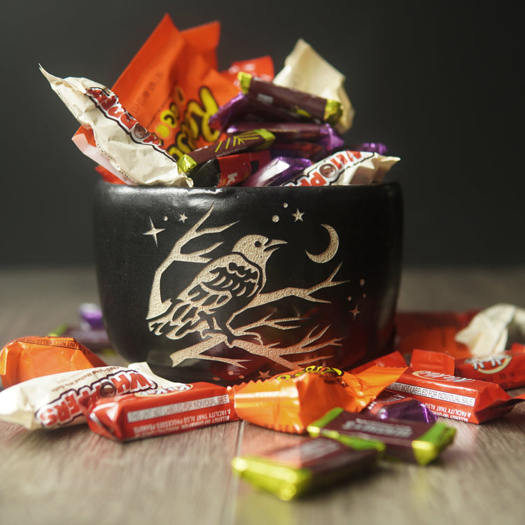 A black pottery bowl overflowing with Halloween candy. A graphic is engraved on the bowl: a crow perched in a tree looking at the crescent moon and stars.