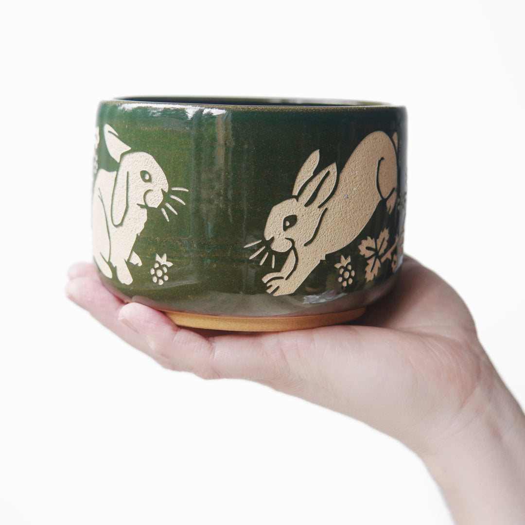 Pine green glazed pottery bowl, engraved with a lop-eared rabbit on the left and a rabbit hopping on the right. There are blackberries near the rabbits' feet. A hand is holding the bowl in the air.