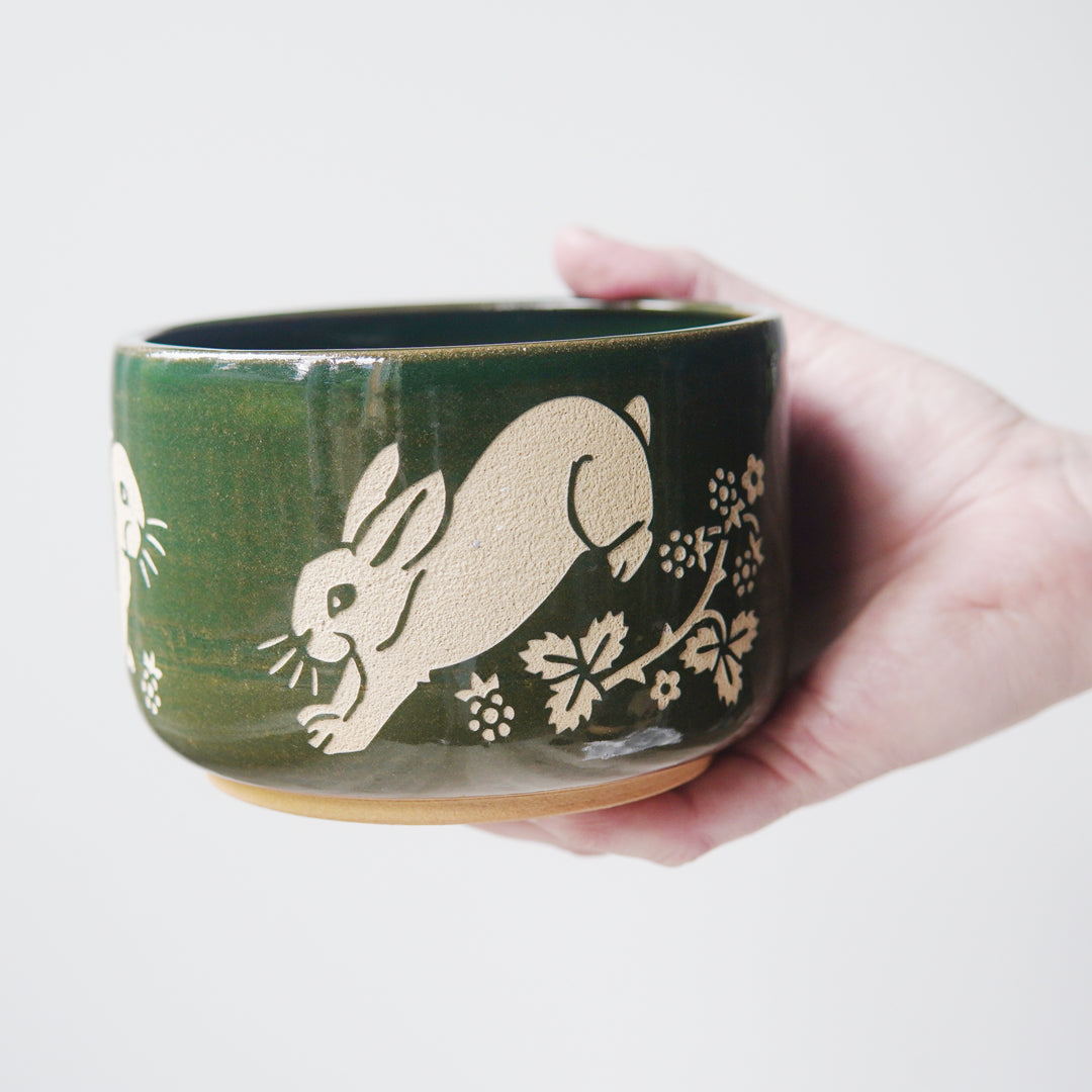 Pine green glazed pottery bowl, engraved with a rabbit hopping. There are blackberries and a thorny plant near the rabbits' feet. A hand is holding the bowl.