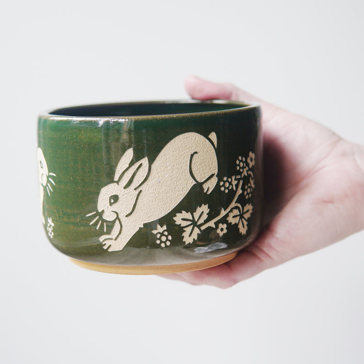 Pine green glazed pottery bowl, engraved with a rabbit hopping. There are blackberries and a thorny plant near the rabbits' feet. A hand is holding the bowl.