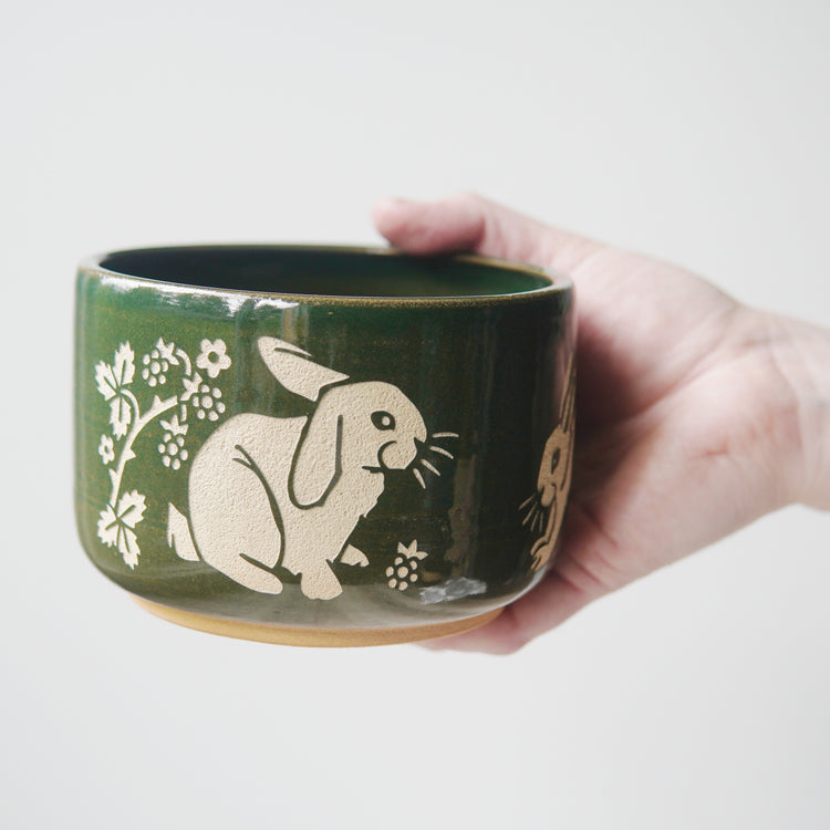 Pine green glazed pottery bowl, engraved with a lop-eared rabbit. There are blackberries and a thorny plant near around the rabbit. A hand is holding the bowl.