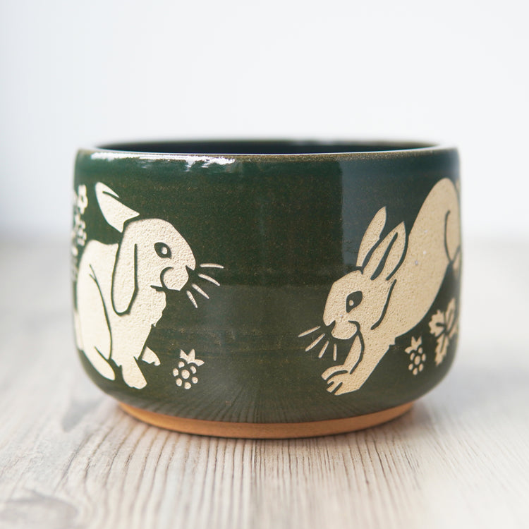Pine green glazed pottery bowl, engraved with a lop-eared rabbit on the left and a rabbit hopping on the right. There are blackberries near the rabbits' feet.