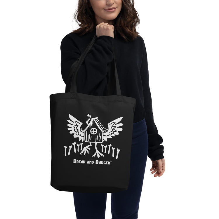 Person holding a black tote bag with a white Baba Yaga's hut graphic and text on a white background