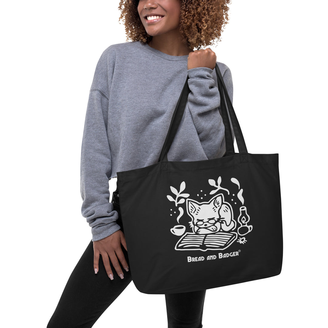 Woman holding a black tote bag with a white cat illustration and 'Bread and Badger' branding on a white background. The cat is sleeping in front of an open book, next to a steaming mug and lantern.