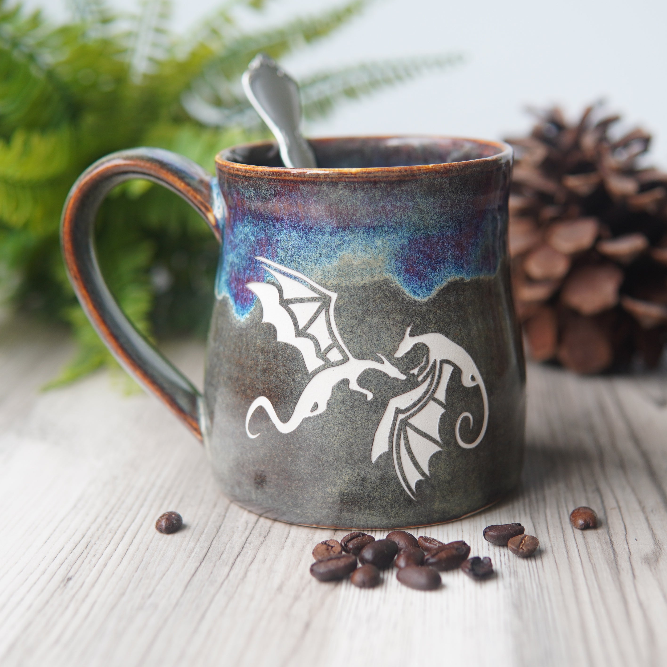 winged dragons engraved on a mug next to coffee beans, a fern, and pinecone