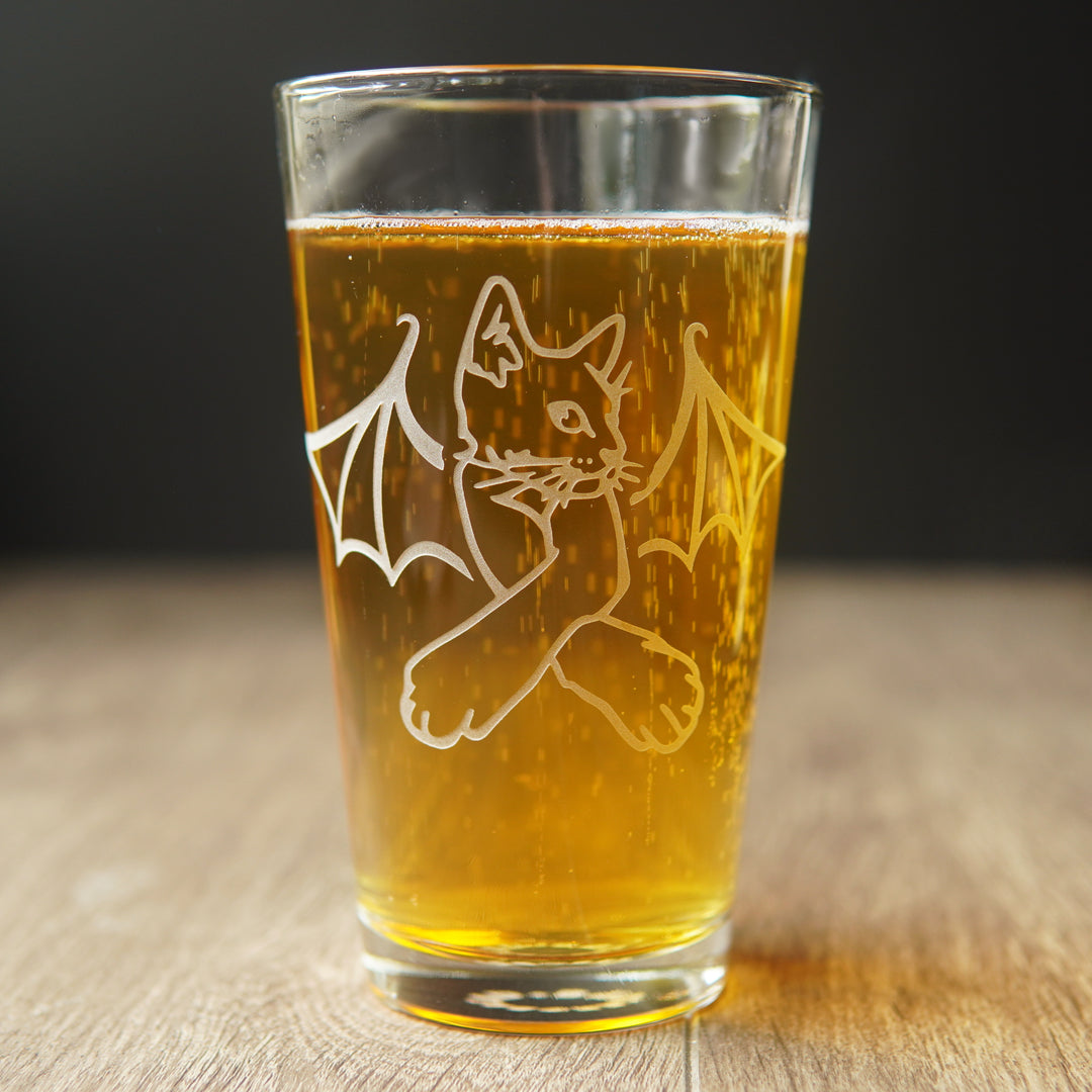 pint glass full of amber beer, engraved with a cat with bat wings