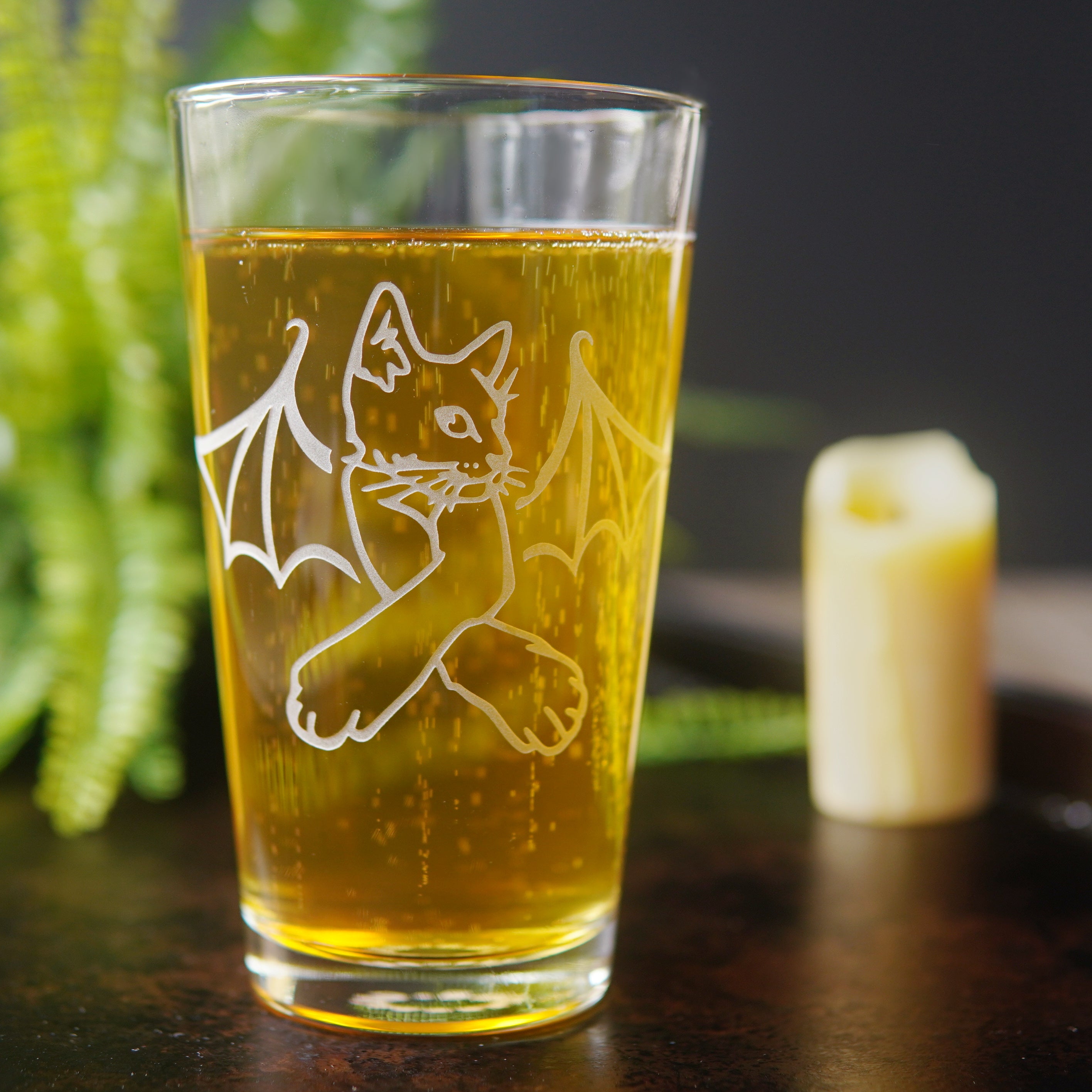 pint glass full of amber beer, engraved with a cat with bat wings