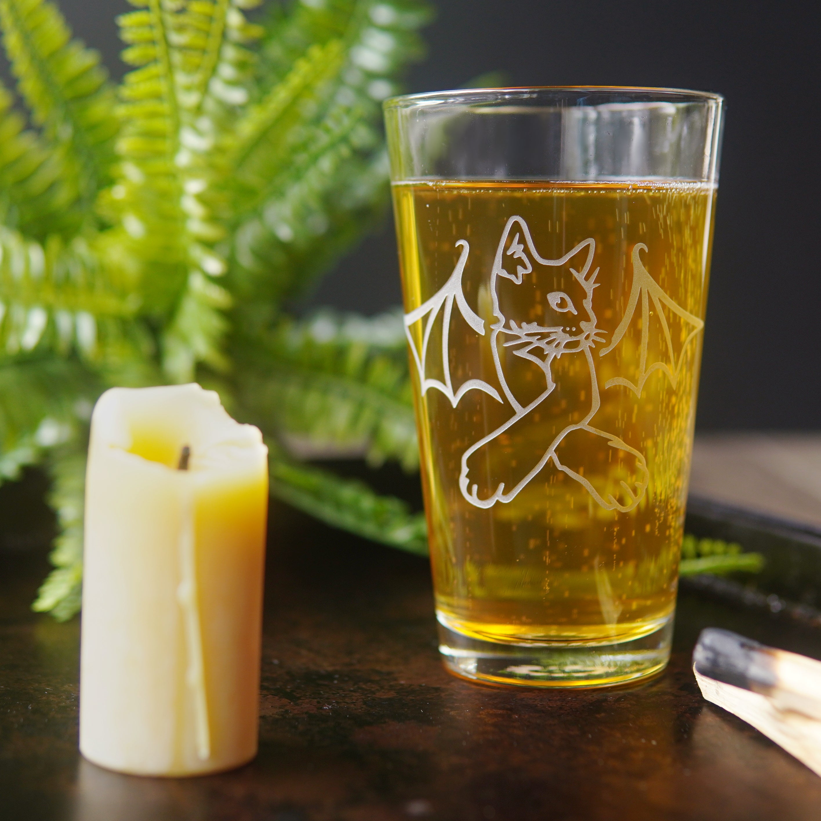 pint glass full of beer, engraved with a cat with bat wings, next to a candle and fern