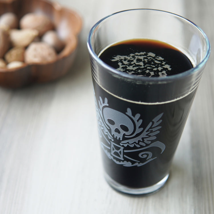 pint glass full of dark beer, engraved with a winged skull, scythes, and hourglass