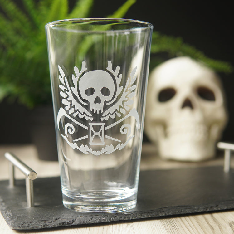 pint glass engraved with a winged skull, scythes, and hourglass. A human skull and fern are in the background