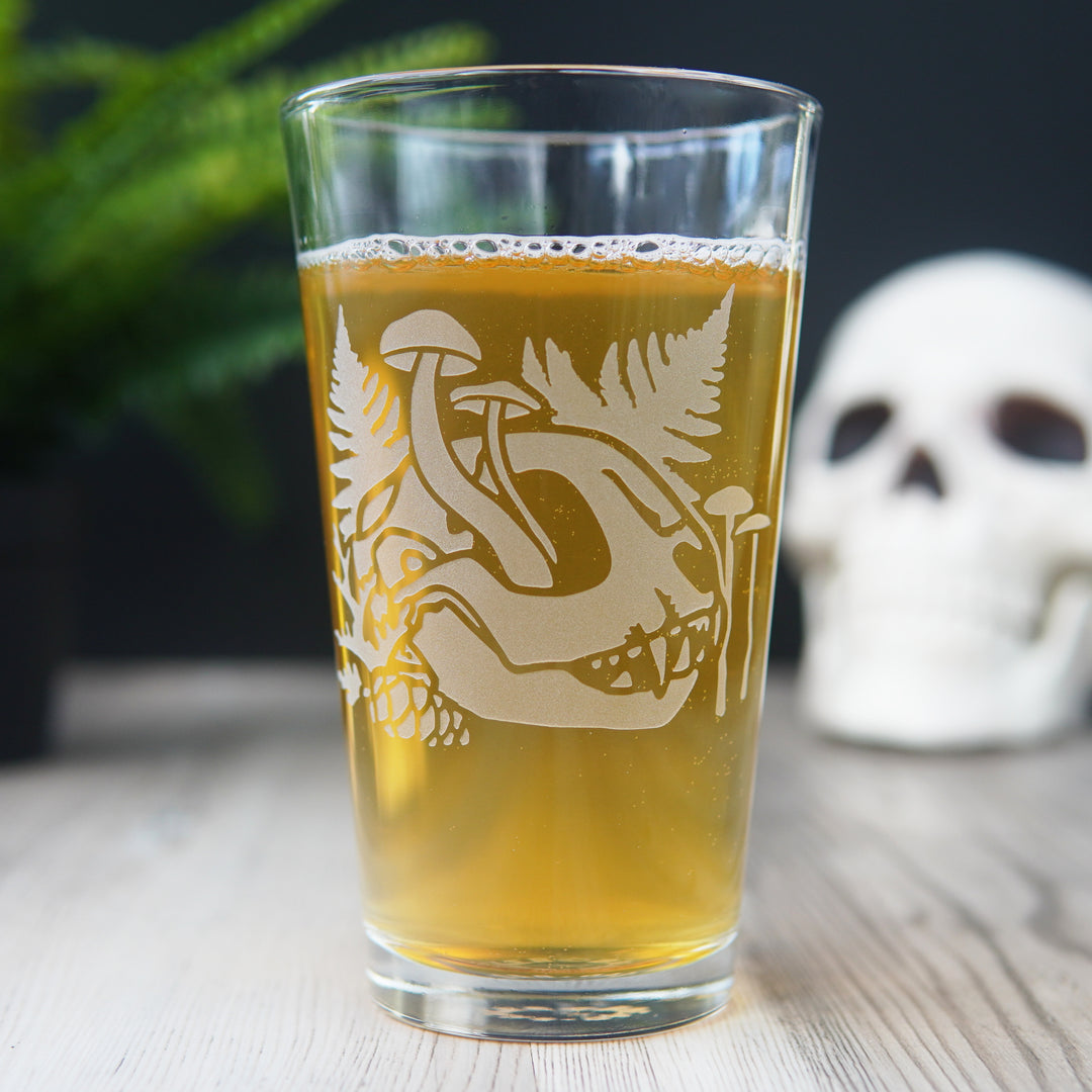 pint glass full of beer engraved with a cat skull, ferns, and mushrooms growing through it
