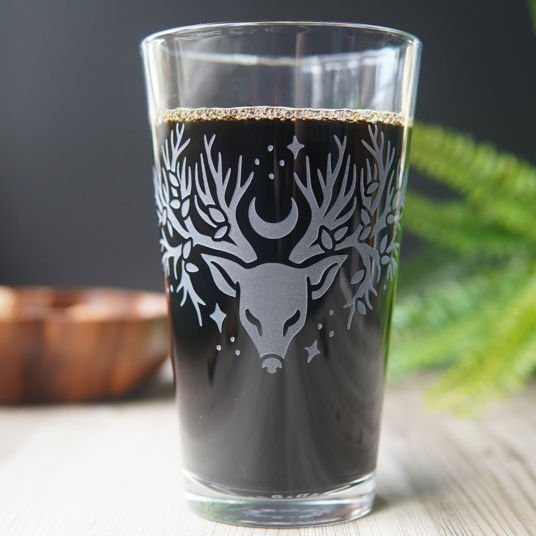 pint glass full of beer, etched with a deer head whose antlers are tree branches with a smattering of leaves, stars, and a sideways crescent moon