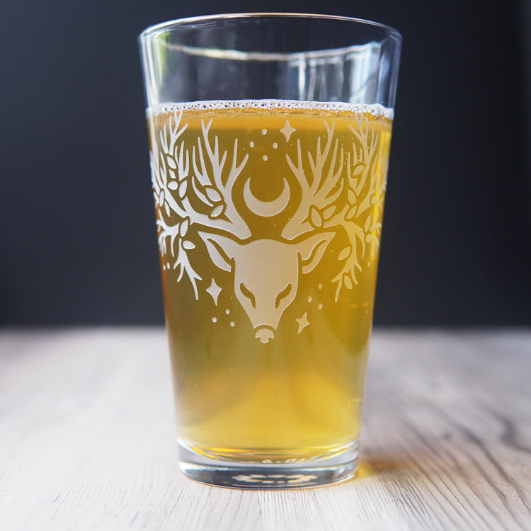 pint glass full of beer, etched with a deer head whose antlers are tree branches with a smattering of leaves, stars, and a sideways crescent moon