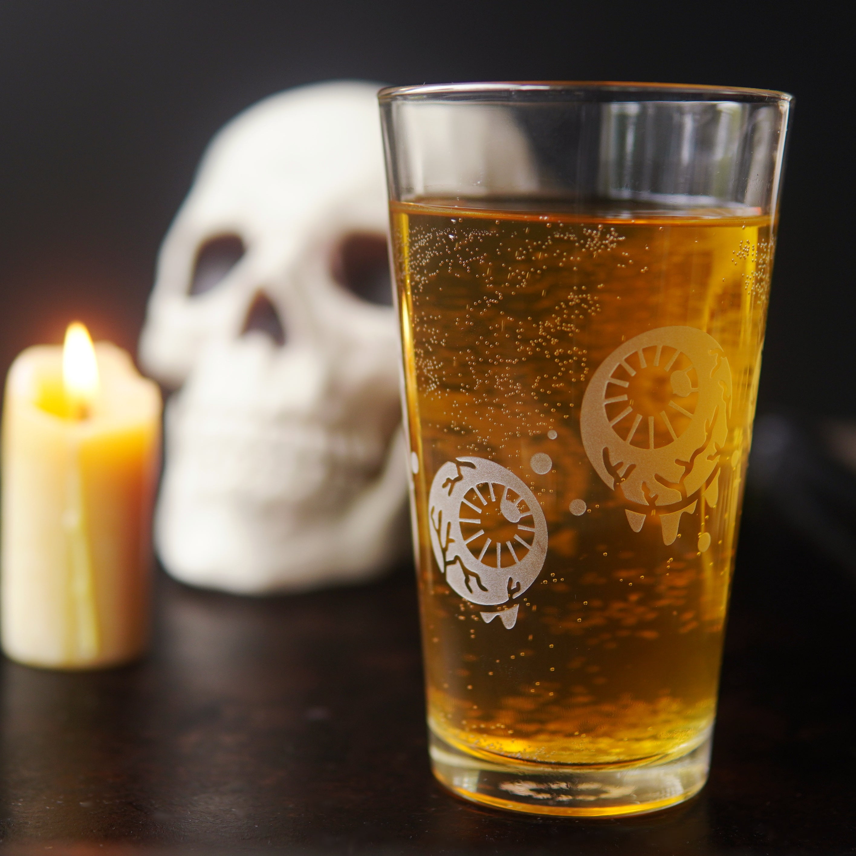 Pint glass engraved with dripping, veiny eyeballs and bubbles, full of beer and sitting next to a lit candle and human skull