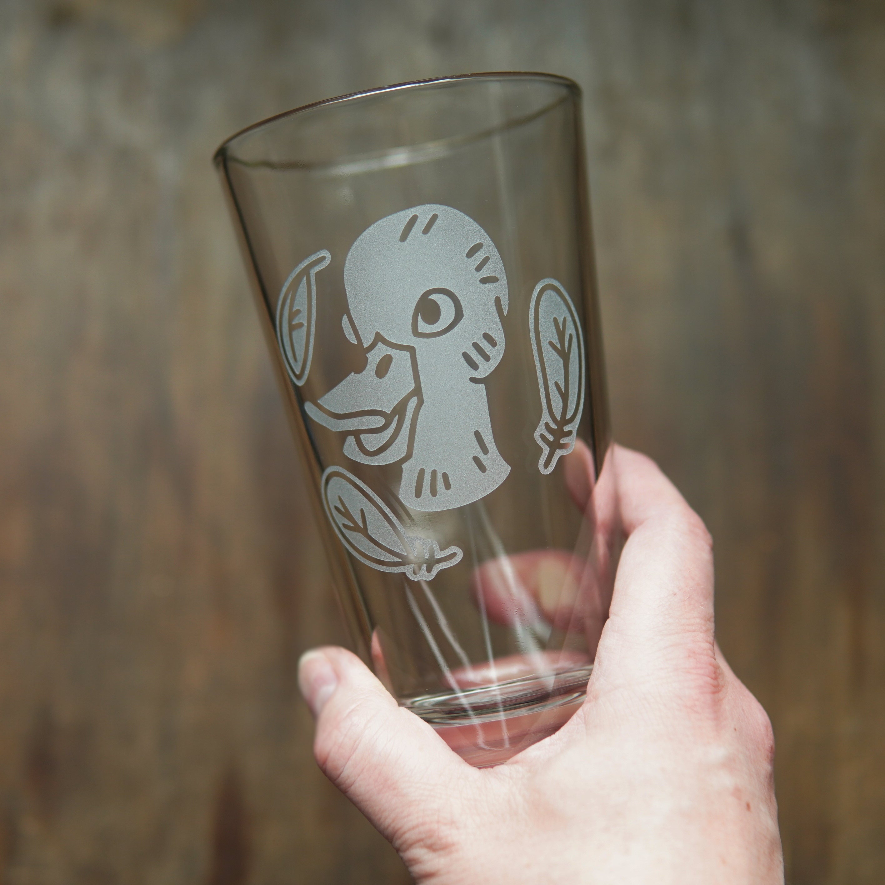 pint glass etched with a duck head, surrounded by three feathers, held by a hand