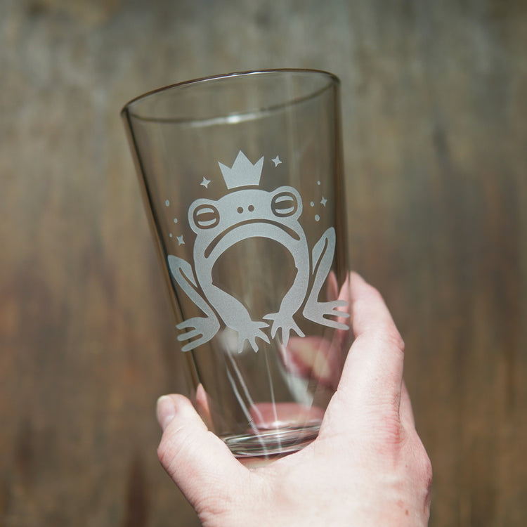 Pint glass etched with a cartoonish frog prince wearing a crown, held by a hand