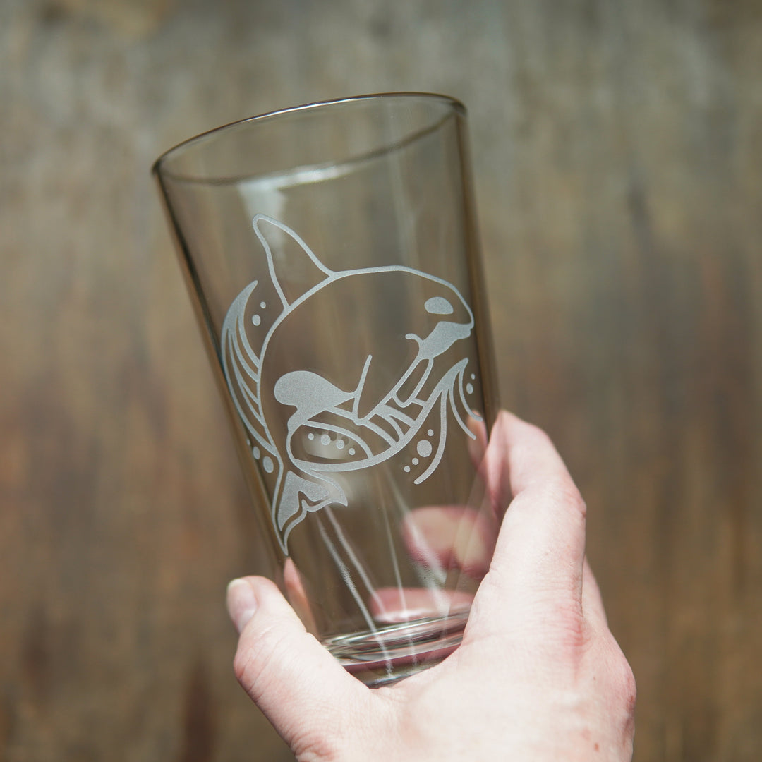 Orca whale and a stylized wave etched on a pint glass, held by a hand