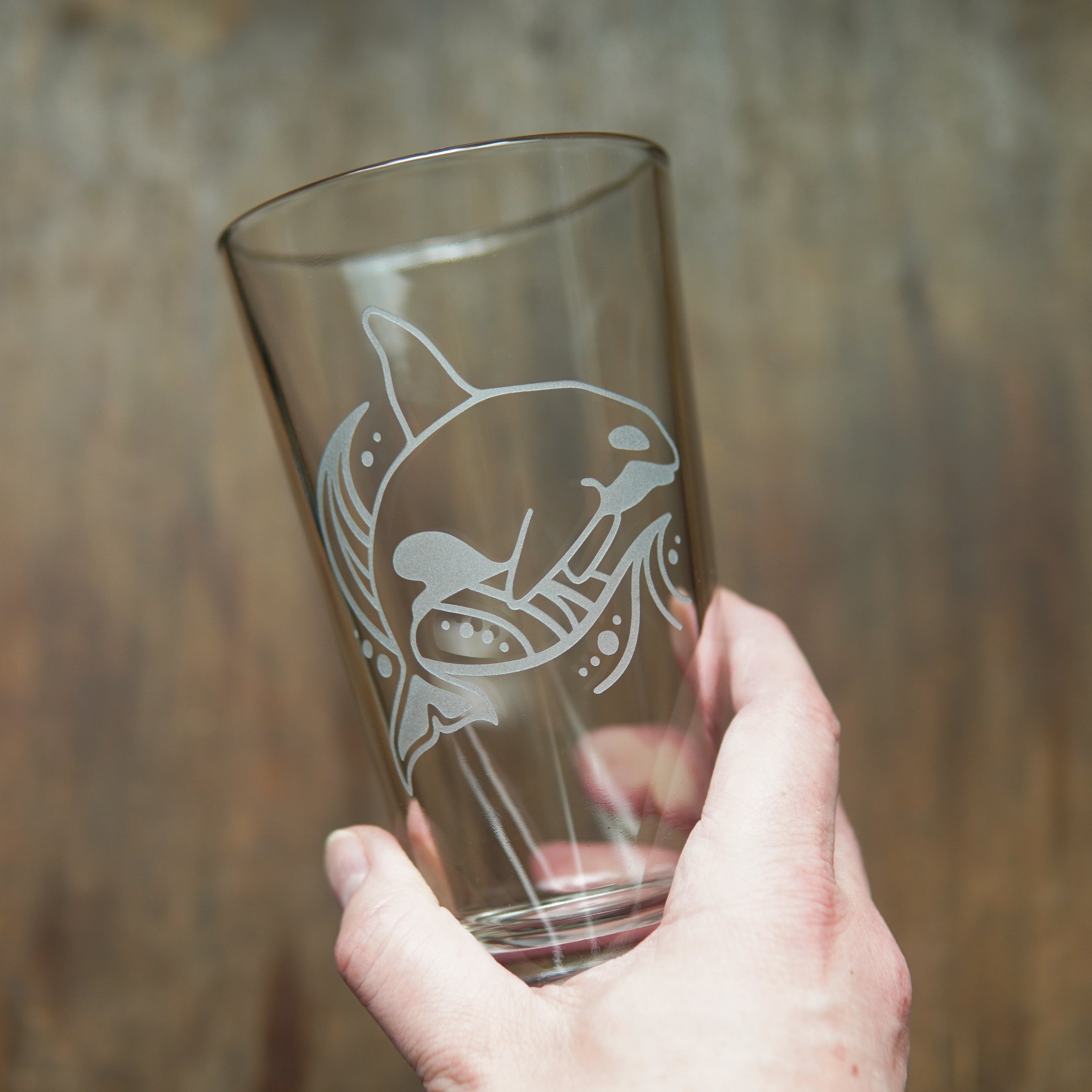 Orca whale and a stylized wave etched on a pint glass, held by a hand