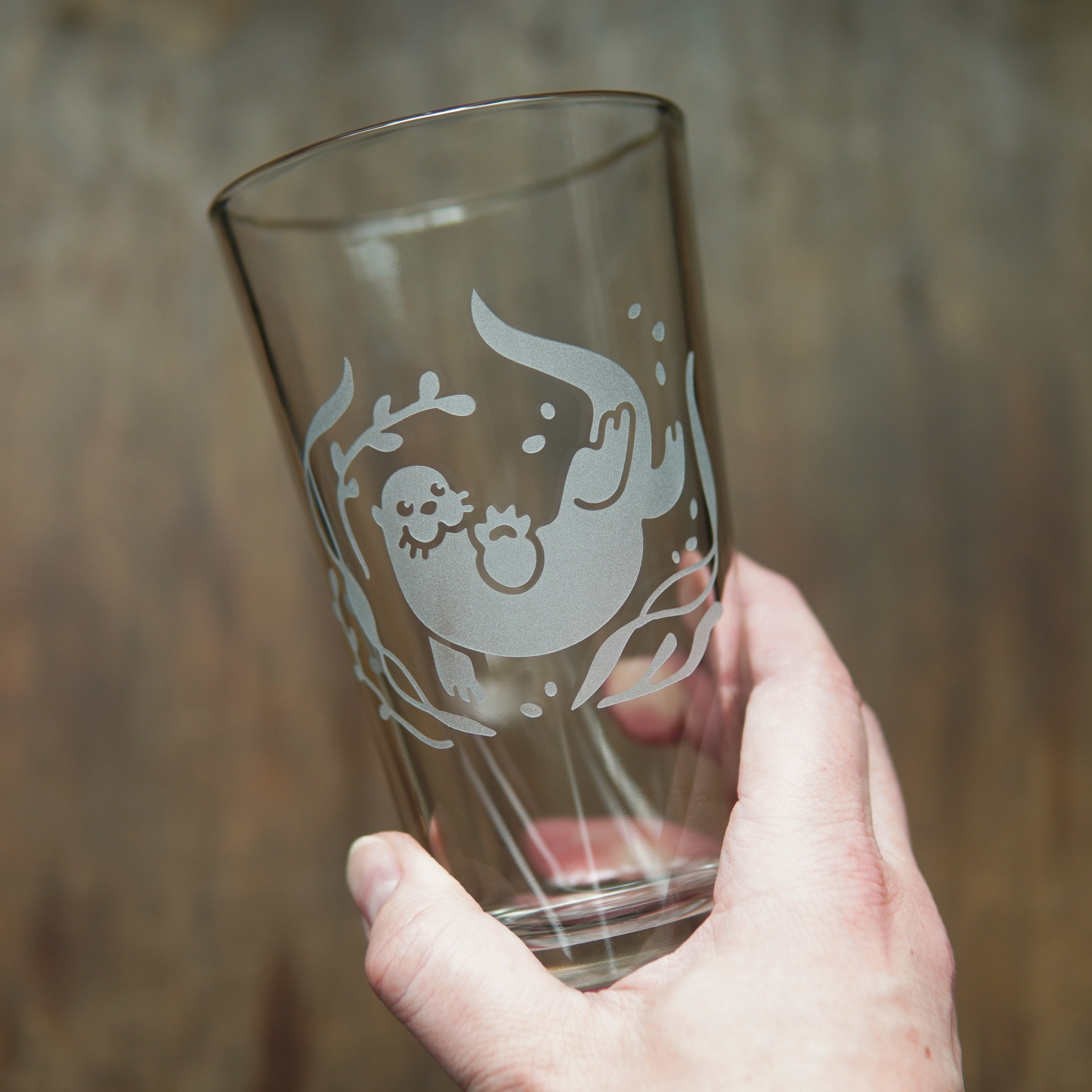 river otter swimming through kelp etched onto a pint glass, held by a hand