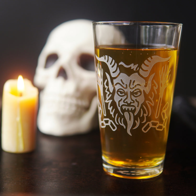 etched Krampus pint glass full of beer, next to a lit candle and human skull