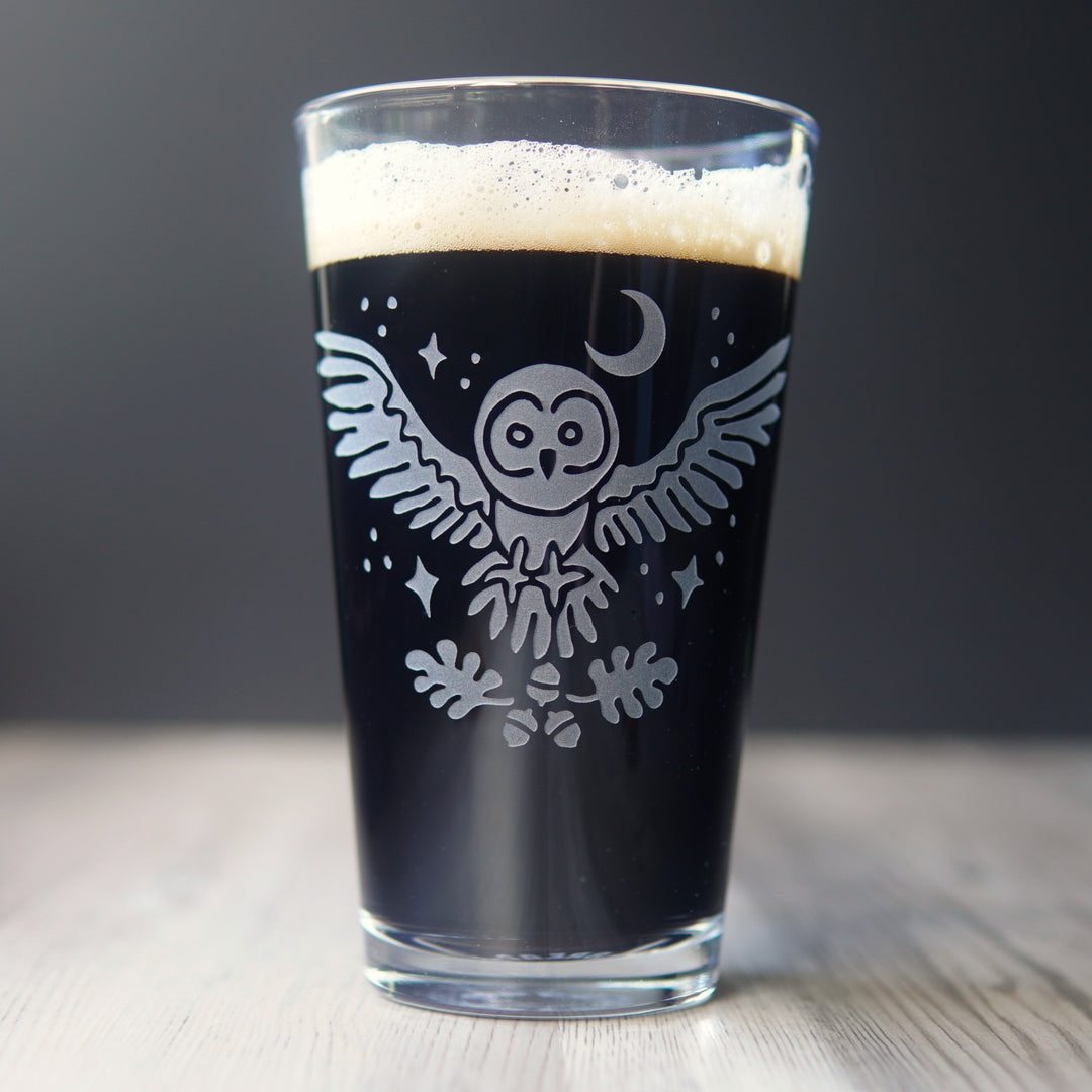 etched pint glass with a flying great grey owl surrounded by stars. A crescent moon hangs above the owl, and oak leaves and acorns float below its open claws