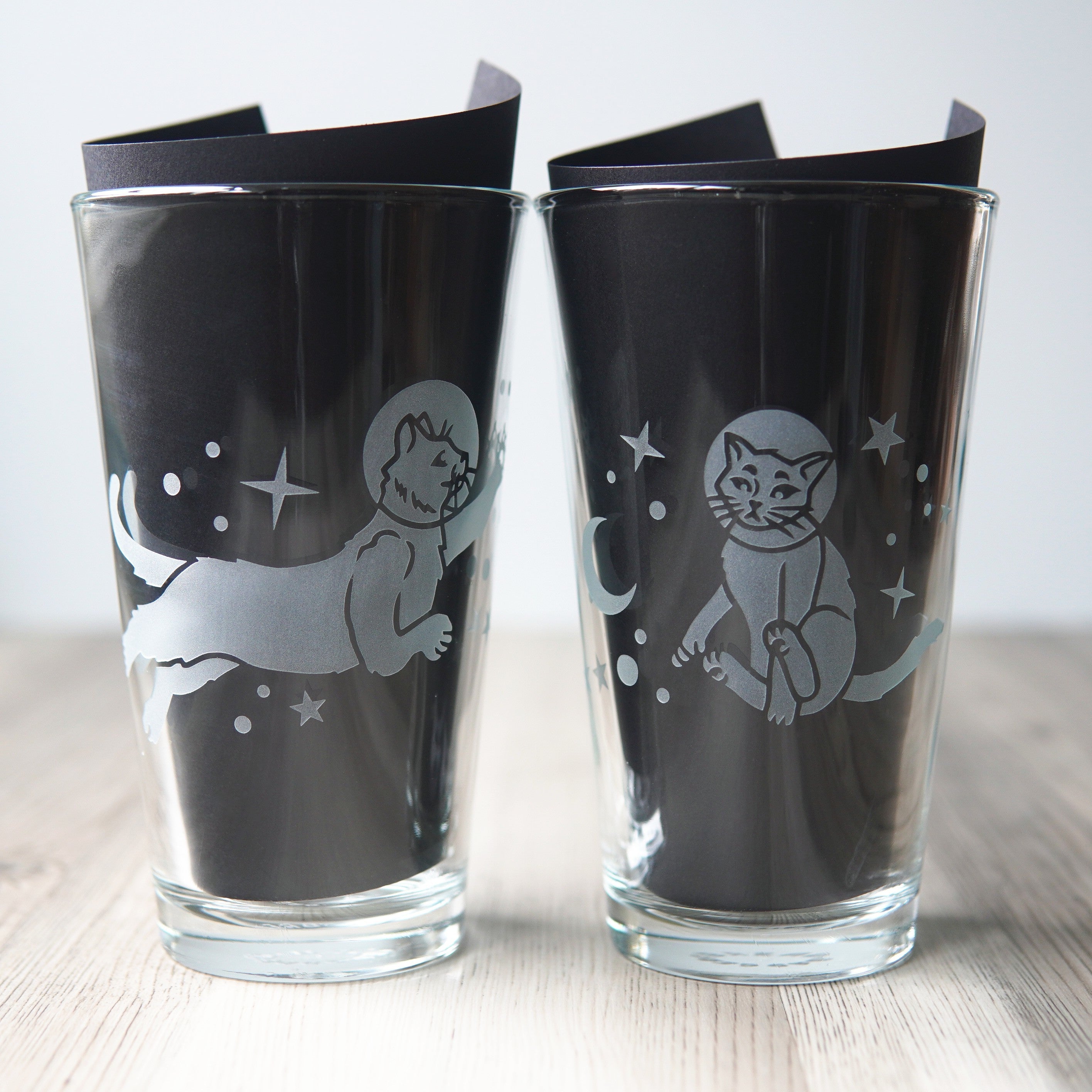 pair of pint glasses engraved with space cats. Black paper is in each glass, to create a background for the clear glass so the etching can be seen