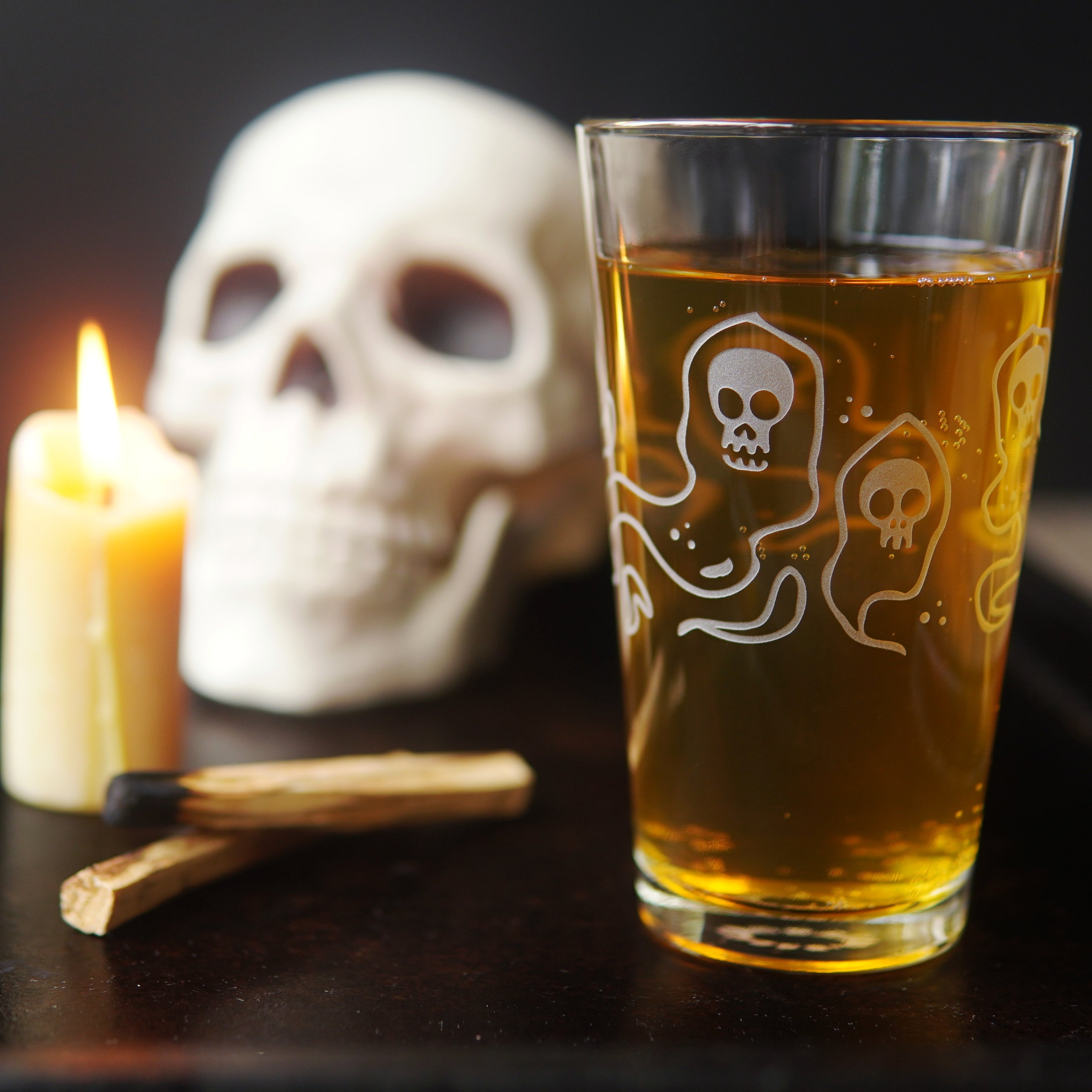 pint glass etched all over with ghosts with skull faces, full of amber beer, next to a lit candle, palo santo, and a plastic skull