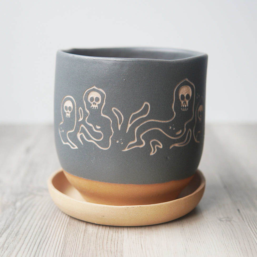 Gray ceramic pot with white ghost skull design on a tan saucer against a light background