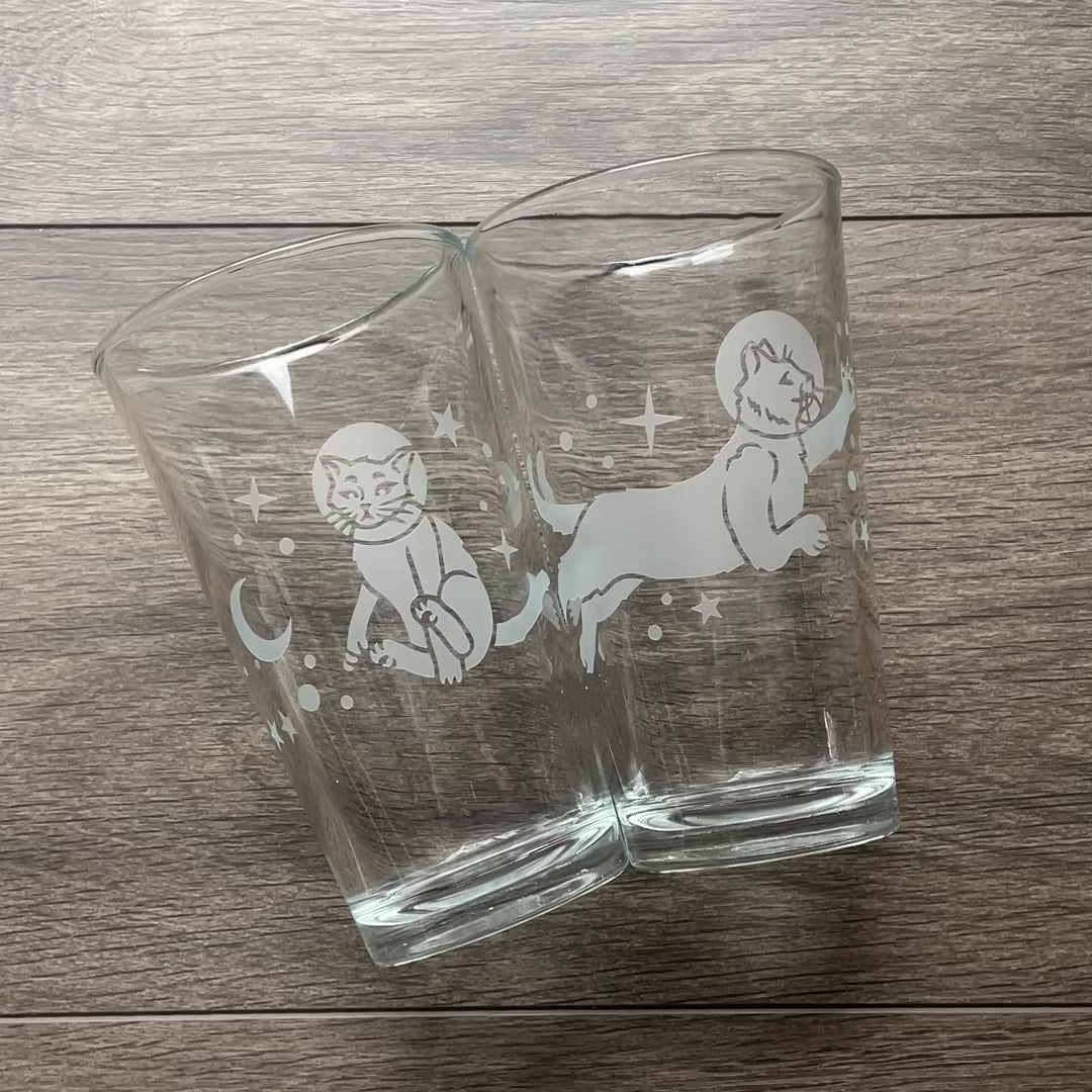 video comparing pint glasses with space cats engraved on them. There is a set of two, one with each cat design, and a third glass with both cat designs wrapping all the way around.