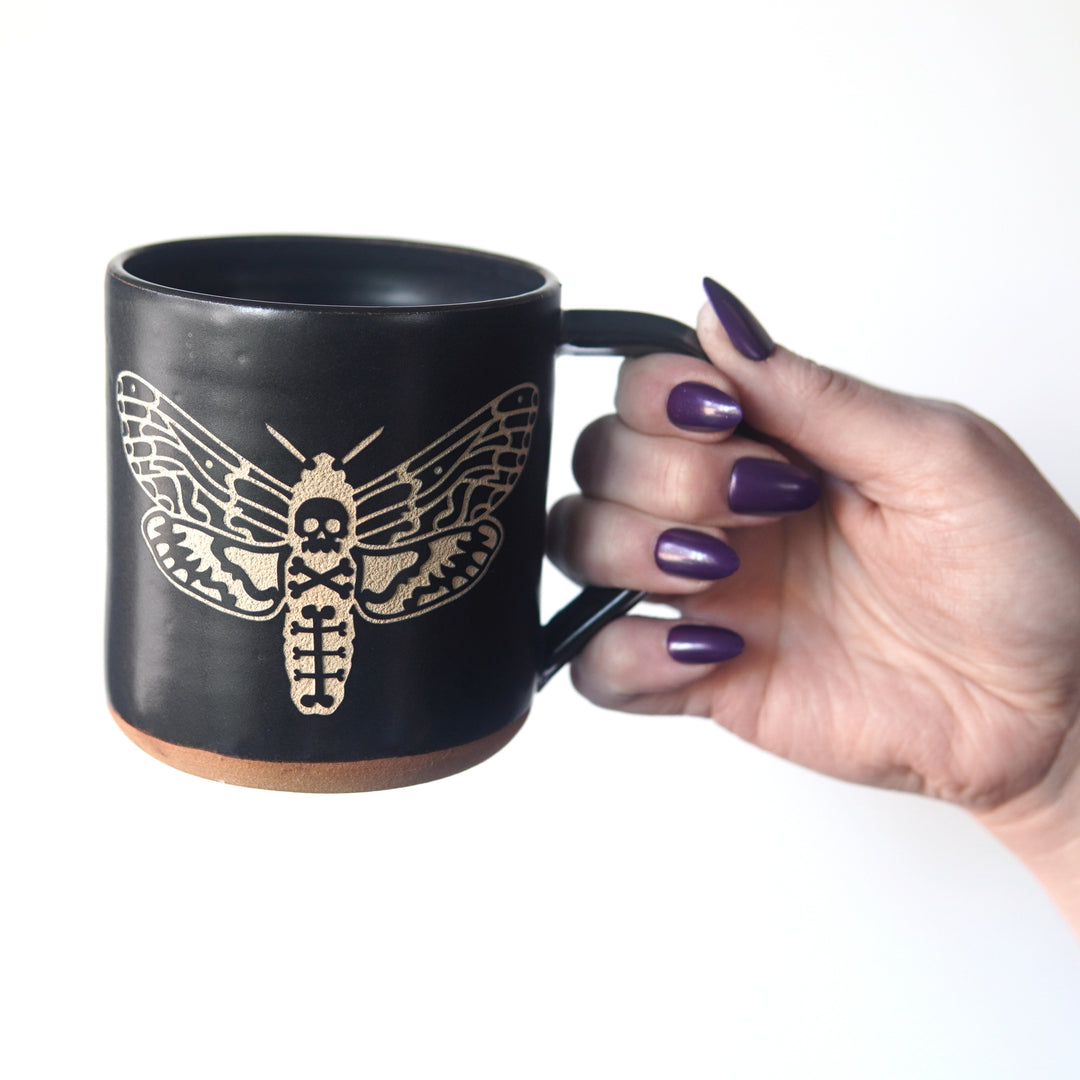 deaths head hawkmoth mug held by the handle. The hand holding the mug has dark purple fingernails.