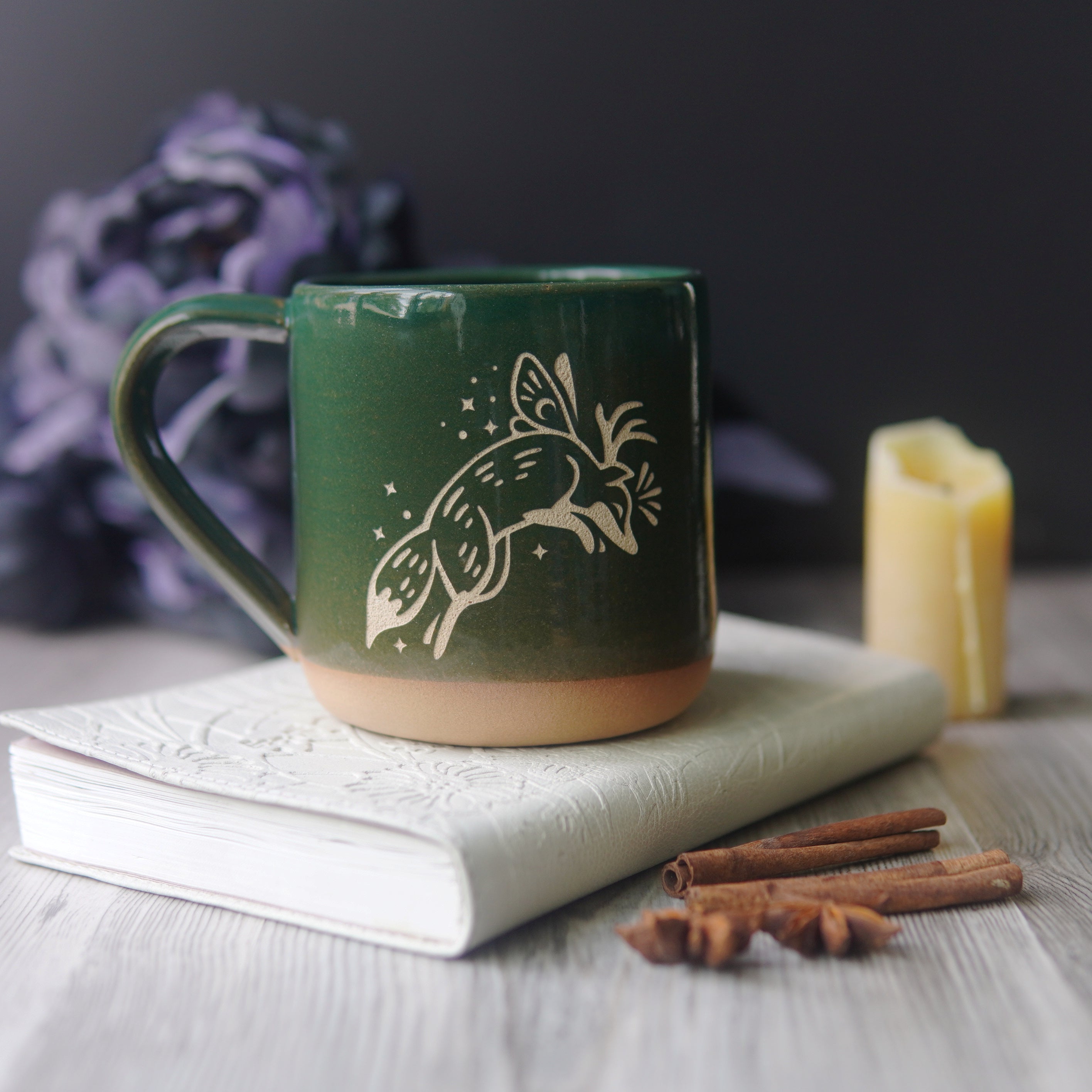 Fox Fairy engraved pottery mug in pine green glaze, with a book, candle, and black flowers