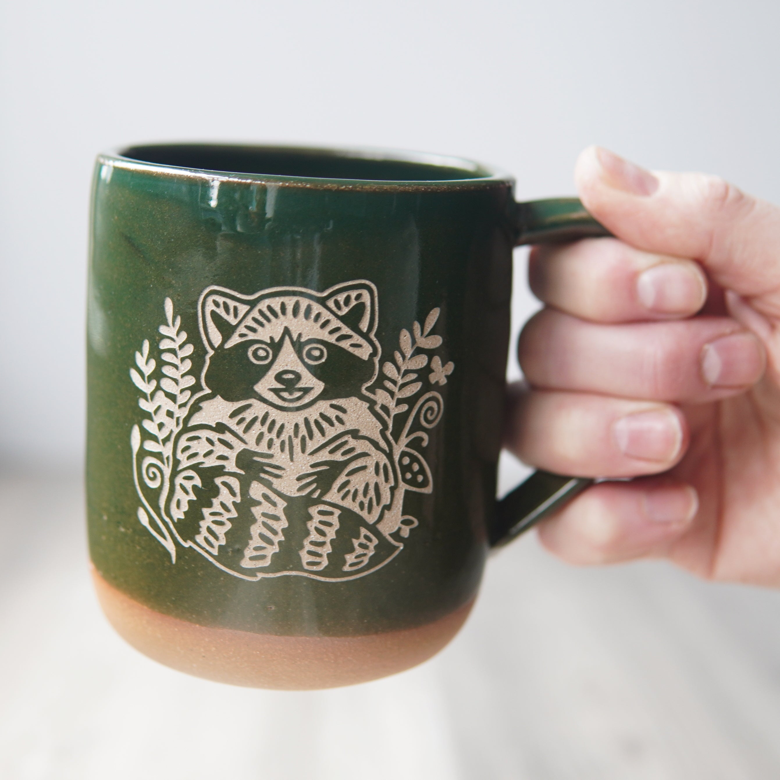 pine green raccoon mug, held by a hand