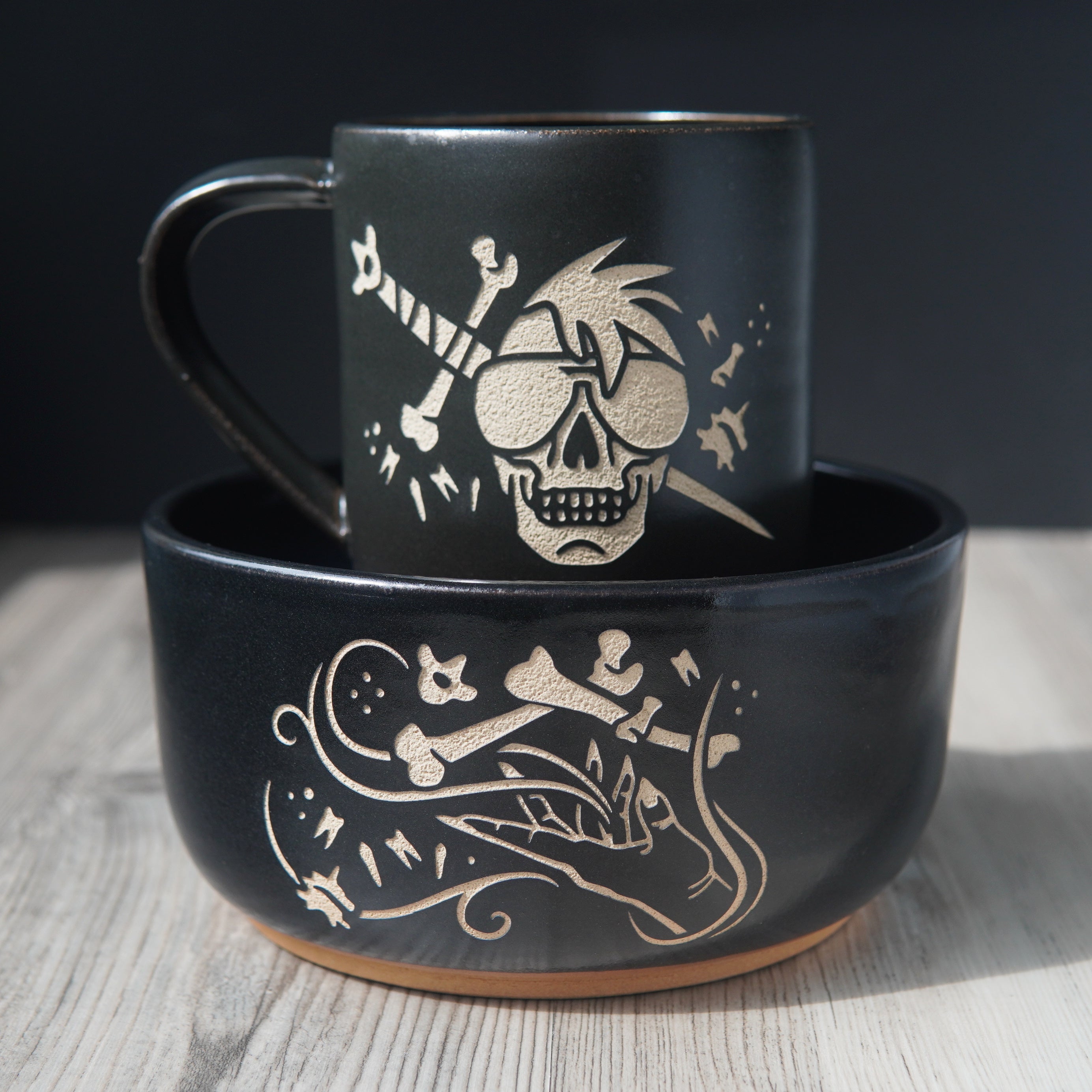 Ramen sized black pottery bowl with a black mug inside it. The bowl has an engraved graphic of a hand levitating human bones and teeth with swirls of magic. The mug is engraved with a skull wearing sunglasses in front of a sword with bones and teeth.