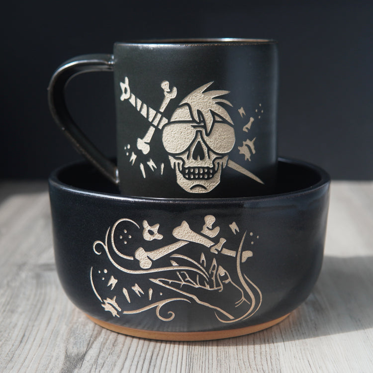 Ramen sized black pottery bowl with a black mug inside it. The bowl has an engraved graphic of a hand levitating human bones and teeth with swirls of magic. The mug is engraved with a skull wearing sunglasses in front of a sword with bones and teeth.