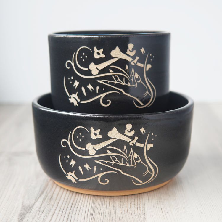 Medium and ramen sized black pottery bowls stacked on each other. The bowls each have an engraved graphic of a hand levitating human bones and teeth with swirls of magic