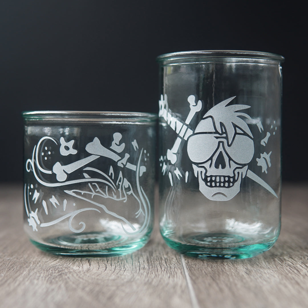 Two straight-sided drinking glasses with a slight green-blue tinge. One is shorter and has a hand and assorted bones on it. The second glass is taller and has a skull with a mohawk wearing sunglasses with a sword and bones behind it.