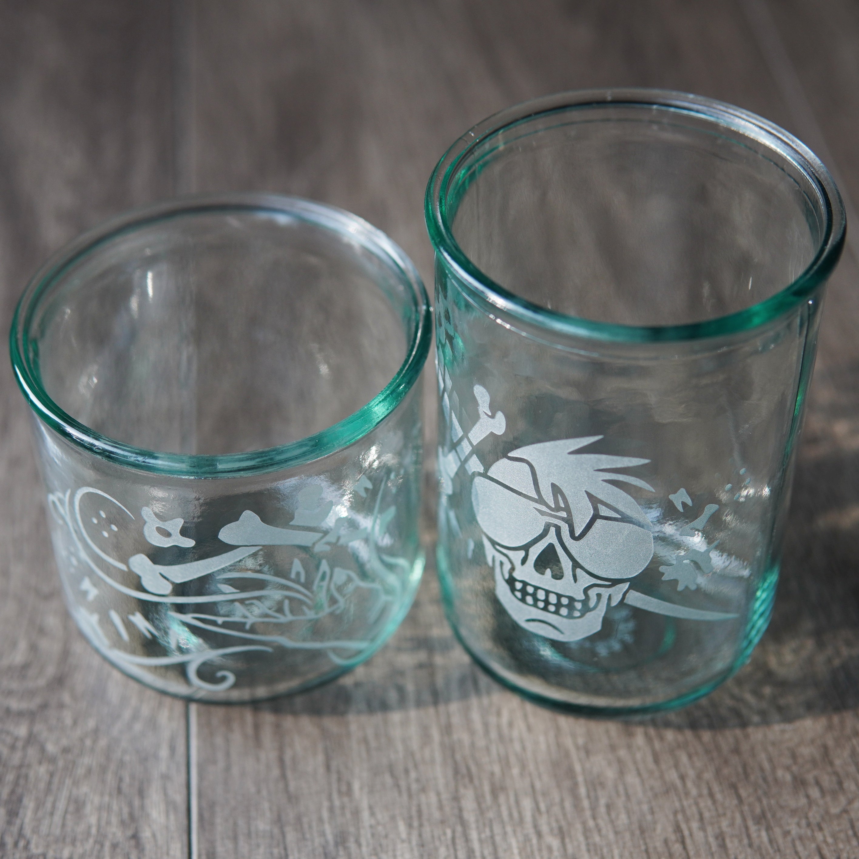 Two straight-sided drinking glasses with a slight green-blue tinge, viewed slightly from above. One is shorter and has a hand and assorted bones on it. The second glass is taller and has a skull with a mohawk wearing sunglasses with a sword and bones behind it.